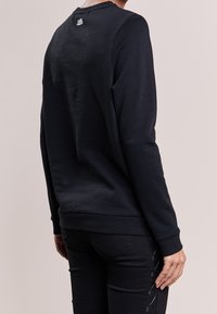 Persona che indossa una felpa nera a maniche lunghe con un piccolo logo bianco "Karl Lagerfeld" sulla parte superiore della schiena, abbinata a pantaloni neri.