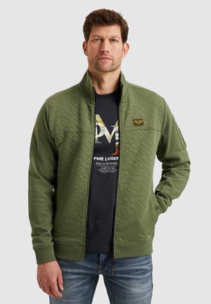 Olijfgroene, gebolde ritsjack met hoge kraag, geribde manchetten en een logo-patch op de borst, gedragen over een zwart T-shirt.