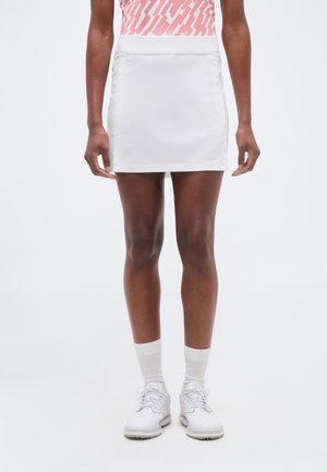 Persoană purtând o fustă sport albă, șosete albe, adidași albi și un top roz cu model, stând pe un fundal simplu.