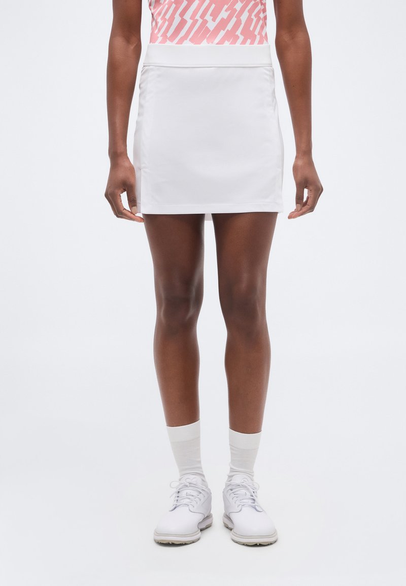 Άτομο που φοράει λευκή αθλητική φούστα, λευκές κάλτσες, λευκά αθλητικά παπούτσια και ροζ τοπ με σχέδια, στέκεται μπροστά σε απλό φόντο.