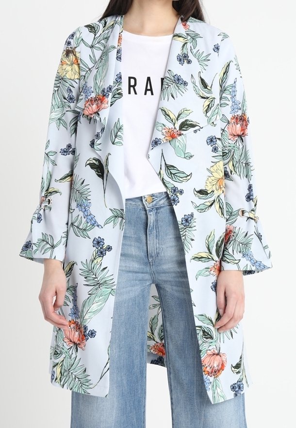Femme portant un manteau fleuri bleu clair par-dessus un T-shirt blanc à motif graphique et un jean taille haute bleu clair, debout devant un fond uni.