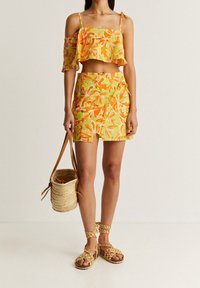 Geel en groene bloemen crop top met geribde mouwen en een bijpassende hooggesneden rok. Model draagt een geweven tas en heeft gevlochten sandalen aan.