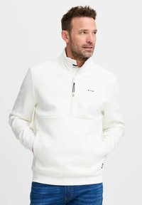 Pullover bianco a metà zip realizzato in morbido tessuto, con tasca frontale, un piccolo logo e polsini a coste. Indossato con jeans in denim blu.