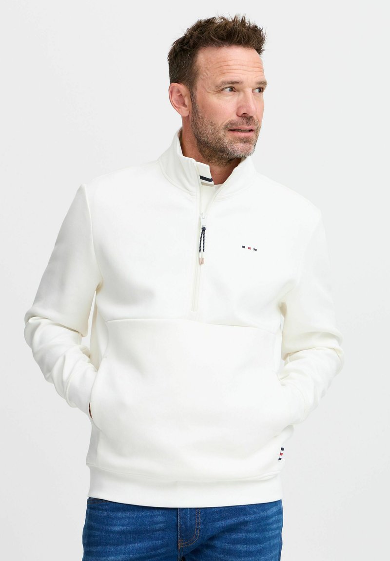 Pullover bianco a metà zip realizzato in morbido tessuto, con tasca frontale, un piccolo logo e polsini a coste. Indossato con jeans in denim blu.