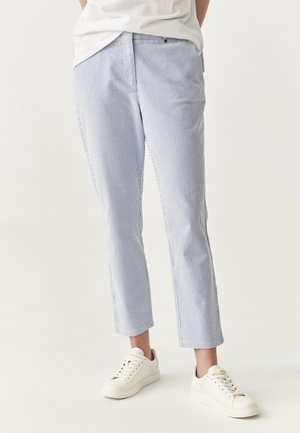 Pantalons rayés bleus et blancs avec une coupe droite, ourlet avec revers et poches avant, assortis à des baskets blanches sur un fond uni.