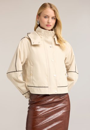 Blonde woman wearing cream jacket with black piping and brown shiny leather skirt, standing against a plain background.