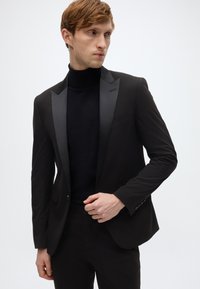Zwart smokingjack met satijnen pieklapels, getailleerde pasvorm, gedragen over een zwarte coltrui. Glad stof, minimale details en een enkele knoopsluiting.