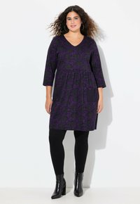 Robe tunique à motif floral violet avec poches, manches trois-quarts et décolleté en V. Associée à des leggings noirs et des bottes à talons.