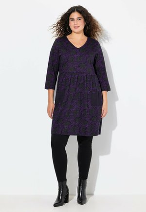Lila blümchenmusterte Tunika-Kleid mit Taschen, Dreiviertelärmeln und V-Ausschnitt. Kombiniert mit schwarzen Leggings und hochhackigen Stiefeln.