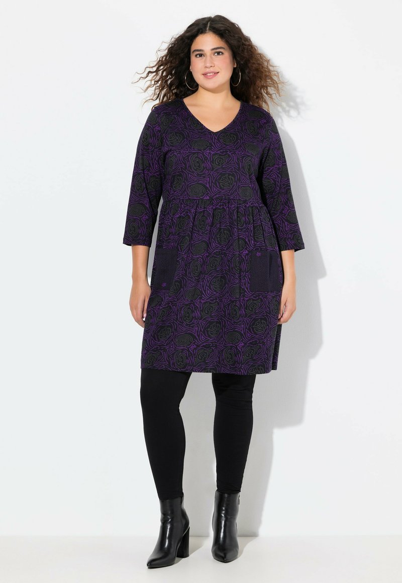 Robe tunique à motif floral violet avec poches, manches trois-quarts et décolleté en V. Associée à des leggings noirs et des bottes à talons.