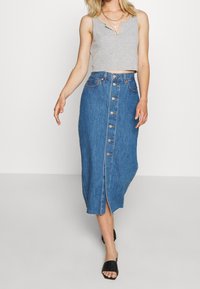 Spódnica midi z denimu z zapięciem na guziki z przodu, bocznymi rozcięciami i niebieskim kolorem. Połączona z szarym, krótko przyciętym topem na ramiączkach i czarnymi sandałami.