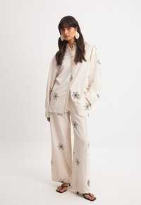 Linne tvådelad outfit bestående av en knapplös skjorta och vida byxor. Beige med svart blommönsterbroderi. Utstickande örhängen och klacksandaler.