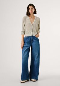 Helle Strickjacke mit V-Ausschnitt und Knopfverschluss, kombiniert mit hochgeschnittenen, weit geschnittenen blauen Jeans. Schlichte weiße Schuhe.