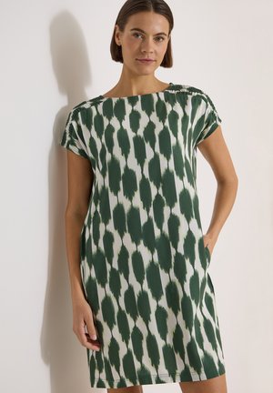 Femme aux cheveux courts et foncés portant une robe blanche à manches courtes avec un motif de coups de pinceau vert foncé, debout contre un mur clair.