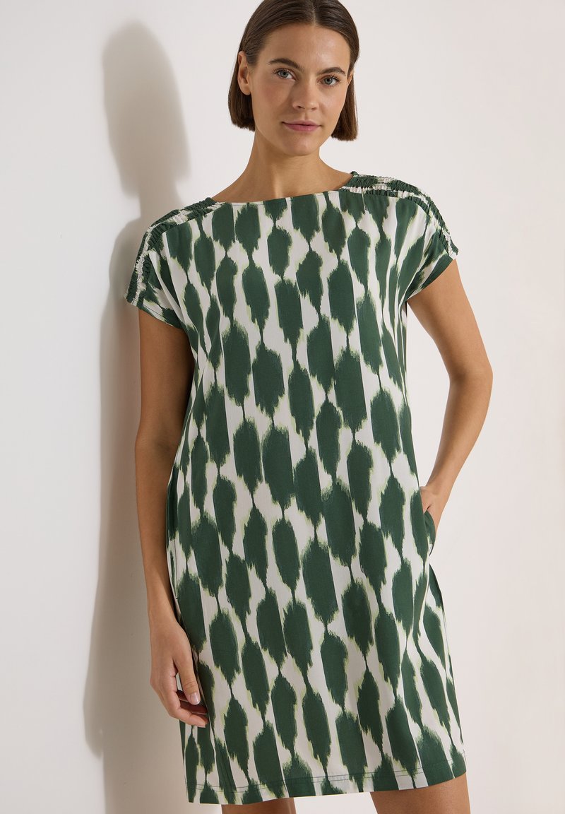 Femme aux cheveux courts et foncés portant une robe blanche à manches courtes avec un motif de coups de pinceau vert foncé, debout contre un mur clair.