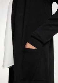 Ruka částečně zasunutá do velké přední kapsy černého pleteného kardiganu přes bílý oděv.