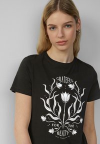 Zwarte katoenen T-shirt met een witte bloemenontwerp en de tekst "Dankbaar voor de Schoonheid" gecentreerd op de voorkant. Korte mouwen.