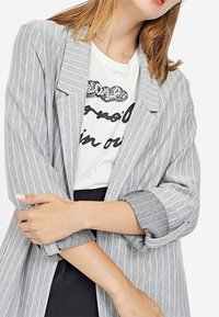Vrouw met lichtbruin haar draagt een grijs gestreepte blazer over een wit graphic t-shirt, met gekruiste armen, tegen een effen witte achtergrond.