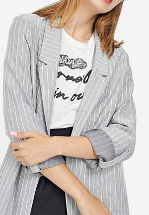 Vrouw met lichtbruin haar draagt een grijs gestreepte blazer over een wit graphic t-shirt, met gekruiste armen, tegen een effen witte achtergrond.