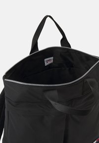 Svart tygshoppingväska med ett dragkedjedetalj, korta handtag och en synlig Tommy Jeans-lapp inuti. Slät textur och minimalistisk design.