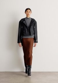 Jaqueta de motoqueiro cropped em couro preto, suéter de gola alta cinza, saia midi castanha e botas pretas até o joelho, sobre um fundo simples.