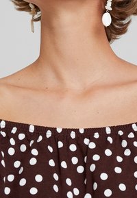 Ruskea olkapäiden yli kulkeva paita, jossa on valkoisia pilkkuja, elastinen kaula-aukko ja teksturoitu kangas. Valkoiset riippuvat korvakorut viimeistelevät asun.