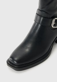 Botine negre din piele, la nivelul gleznei, cu vârful rotund, toc jos și un accent de cataramă argintie pe curea. Textură netedă cu detalii minime de cusătură.