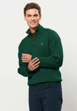 Jeune homme souriant portant un pull vert en maille torsadée avec fermeture éclair quart de zip et un pantalon bleu foncé sur un fond clair uni.