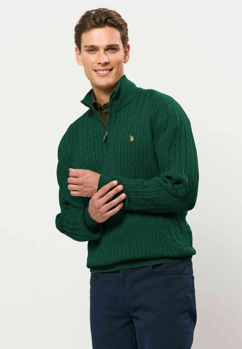Jeune homme souriant portant un pull vert en maille torsadée avec fermeture éclair quart de zip et un pantalon bleu foncé sur un fond clair uni.