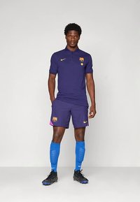 Barcelona fotbollströja i mörklila. Pikétröja med gul Nike-logotyp och klubbemblem, shorts i matchande färg med rosa detaljer, blå strumpor, svarta skor.
