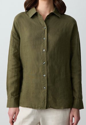 Mujer con una camisa de manga larga color verde oliva con botones y cuello, y pantalones de color claro, de pie frente a un fondo liso.