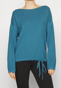 Pull turquoise en tricot doux. Coupe ample avec un col large et des manches longues. Équipé d'un ourlet à cordon avec des accents en métal.