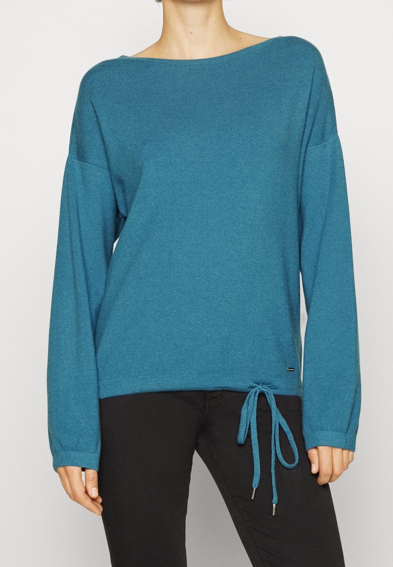 Pull turquoise en tricot doux. Coupe ample avec un col large et des manches longues. Équipé d'un ourlet à cordon avec des accents en métal.