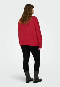 Maglione rosso lavorato a maglia con una vestibilità ampia, spalle scese e texture a coste, abbinato a pantaloni neri aderenti e stivaletti neri alla caviglia.