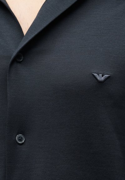 Κοντινή λήψη μαύρου πουκάμισου με κουμπιά και μικρό κεντημένο λογότυπο πουλιού στο αριστερό στήθος, με ορατό μέρος του λαιμού.