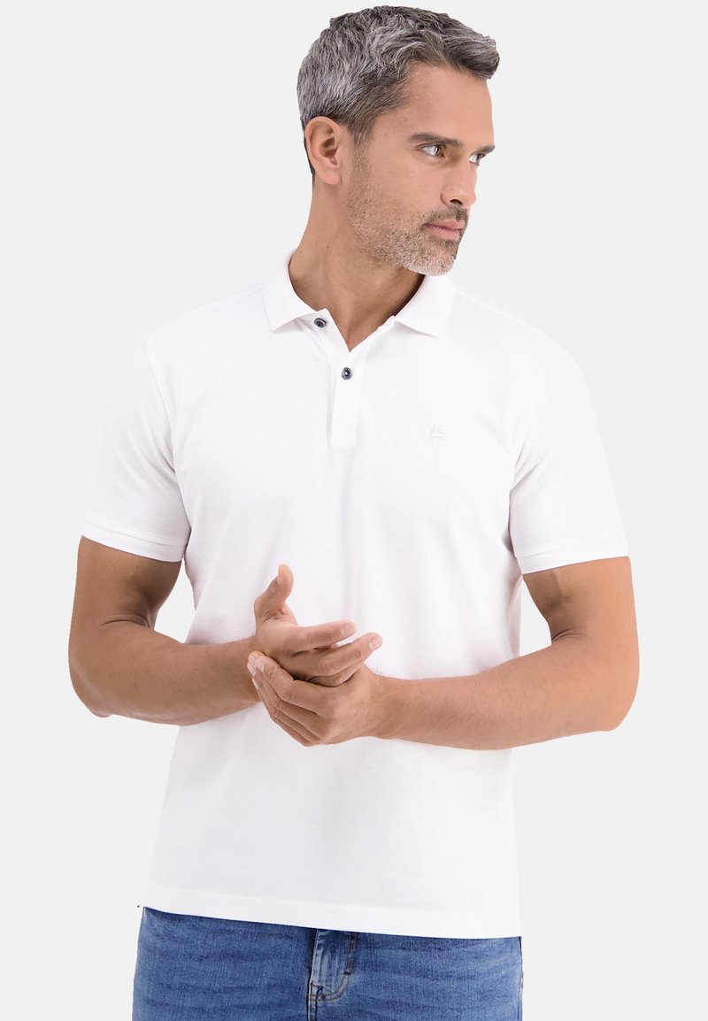 Mann mit kurzem grauem Haar, der ein weißes Polohemd und blaue Jeans trägt und mit verschränkten Händen vor sich nach rechts schaut.