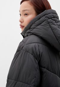 Czarna kurtka puchowa z wysokim kołnierzem i kapturem. Posiada pikowaną strukturę i smukły, aerodynamiczny kształt. Gładkie wykończenie powierzchni.