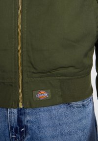 Olivgrön jacka med hög krage, gulddragkedja, ribbad midja och en Dickies-logotyp, kombinerad med blå denimjeans.