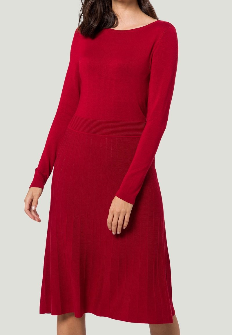 Femme portant une robe en maille rouge à manches longues avec une taille cintrée et une jupe côtelée sur un fond clair uni.