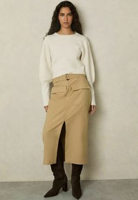 Beige Rock mit vorderem Schlitz und zwei großen Vordertaschen, kombiniert mit einem weißen, strukturierten Langarmoberteil und dunkelbraunen, knielangen, hochhackigen Stiefeln.