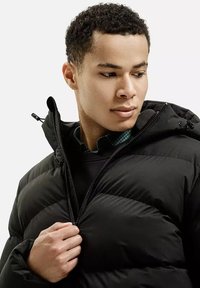 Czarna puchowa kurtka z zapięciem na zamek, z kapturem i elastycznymi mankietami, noszona na ciemnym swetrze i zielonej koszuli w kratę.