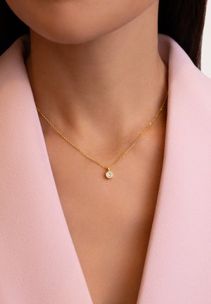 Collar de oro con una cadena fina, que presenta un pequeño colgante redondo engastado con una piedra clara, luciendo sobre un fondo de tela de color rosa claro.