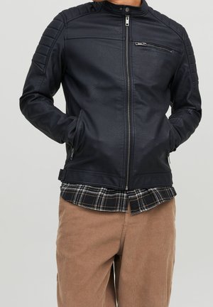 Chaqueta de cuero negra con detalles acanalados en los hombros, cierre frontal con cremallera y dos bolsillos laterales. Llevada sobre una camisa de cuadros y pantalones beige.