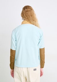 Persoană cu păr lung, ondulat și blond, purtând o jachetă albastră deschisă cu petice maro pe coate și pantaloni crem, întoarsă cu spatele, pe un fundal alb.