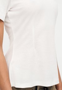 Κλείσιμο σε πρόσωπο που φοράει απλό λευκό κοντομάνικο μπλουζάκι και καφέ παντελόνι μπροστά σε φωτεινό φόντο.