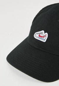 Svart baseballcap laget av bomull, med en hvit brodert sneakerlogo med røde og blå aksenter på forsiden.