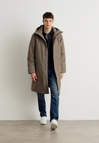 Lang, olivengrønn polstret kåpe med hette, med glidelås og to sidelommer. Bæres over en svart skjorte og blå jeans.