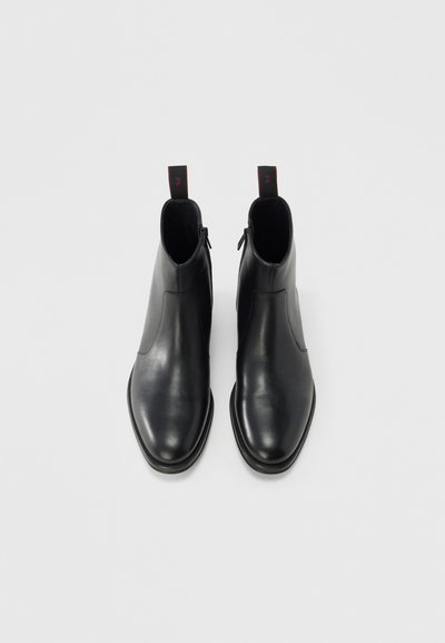 Botines de cuero negro con un acabado elegante y suave, punta afilada y cierre de cremallera, con tiradores rojos en la parte trasera.