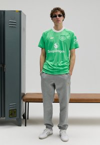 Zöld atlétatrikó fehér díszítésekkel, logóval. Szürke melegítőnadrággal és fehér tornacipővel párosítva, szürke szekrény előtt állva.