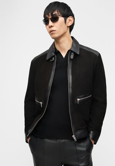 Άνδρας που φοράει μαύρο μπουφάν με φερμουάρ και δερμάτινες λεπτομέρειες, μαύρο ριμπ πουλόβερ με V λαιμόκοψη, μαύρο δερμάτινο παντελόνι και σκούρα γυαλιά ηλίου, με τα χέρια στις τσέπες.